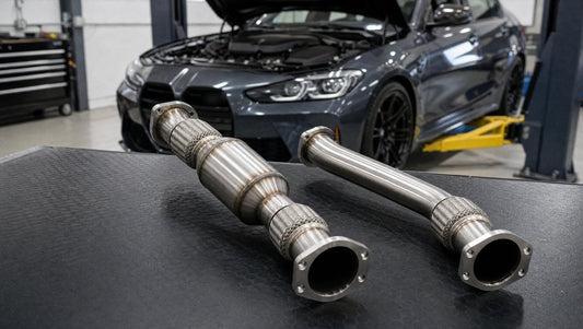 Catted and catless stainless steel turbo downpipes side by side on a workbench, with a BMW sedan on a lift behind