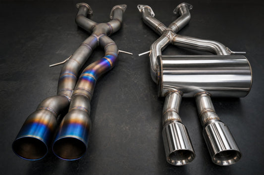 Titanium exhaust with blue and gold heat-tinted pipes next to a polished stainless steel cat-back exhaust with a round muffler on a workshop bench