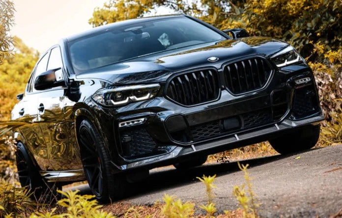 A tilted front view of a black car outside on the road fitted with AUTOTECKNIC PERFORMANCE DRY CARBON FRONT LIP SET - BMW G06 X6 M-SPORT 2023+