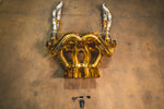 Overhead view of a gold-plated Valvetronic Designs exhaust system for a Lamborghini Aventador, including heat-shielded piping and a remote control module.