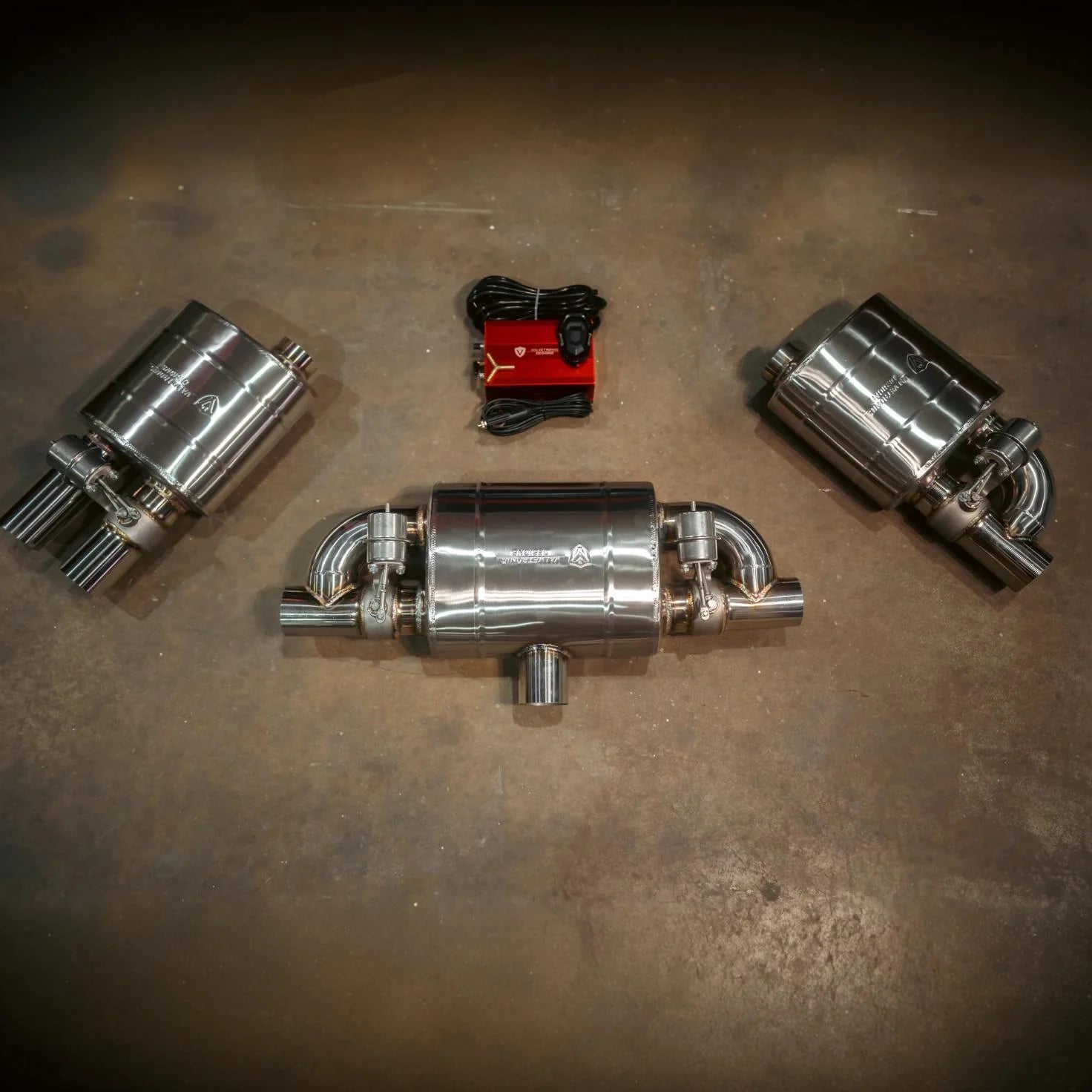 A wide-angle view of a universal valved muffler kit including three polished stainless steel mufflers, a red electronic control box, and wiring harnesses laid out on a concrete floor.