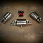 A wide-angle view of a universal valved muffler kit including three polished stainless steel mufflers, a red electronic control box, and wiring harnesses laid out on a concrete floor.