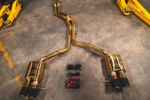 A full view of an anodized gold dual-exit exhaust system laid out on a concrete floor, featuring mid-pipes, two mufflers, carbon fiber exhaust tips, and accompanying wiring harnesses with a control box.
