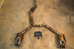 A full view of a titanium cat-back exhaust system with heat-treated gold and purple finishes laid out on a workshop floor, including two valved mufflers and a central control module.
