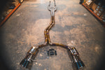 A wide-angle top view of a titanium cat-back exhaust system with a gold-to-purple heat-treated finish, laid out on a concrete floor with its wiring harness and control module.