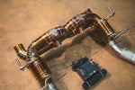 An angled overhead view of the gold-finish valved catback exhaust system showing the intricate TIG welding, dual muffler canisters, and a wireless remote control unit on the floor.