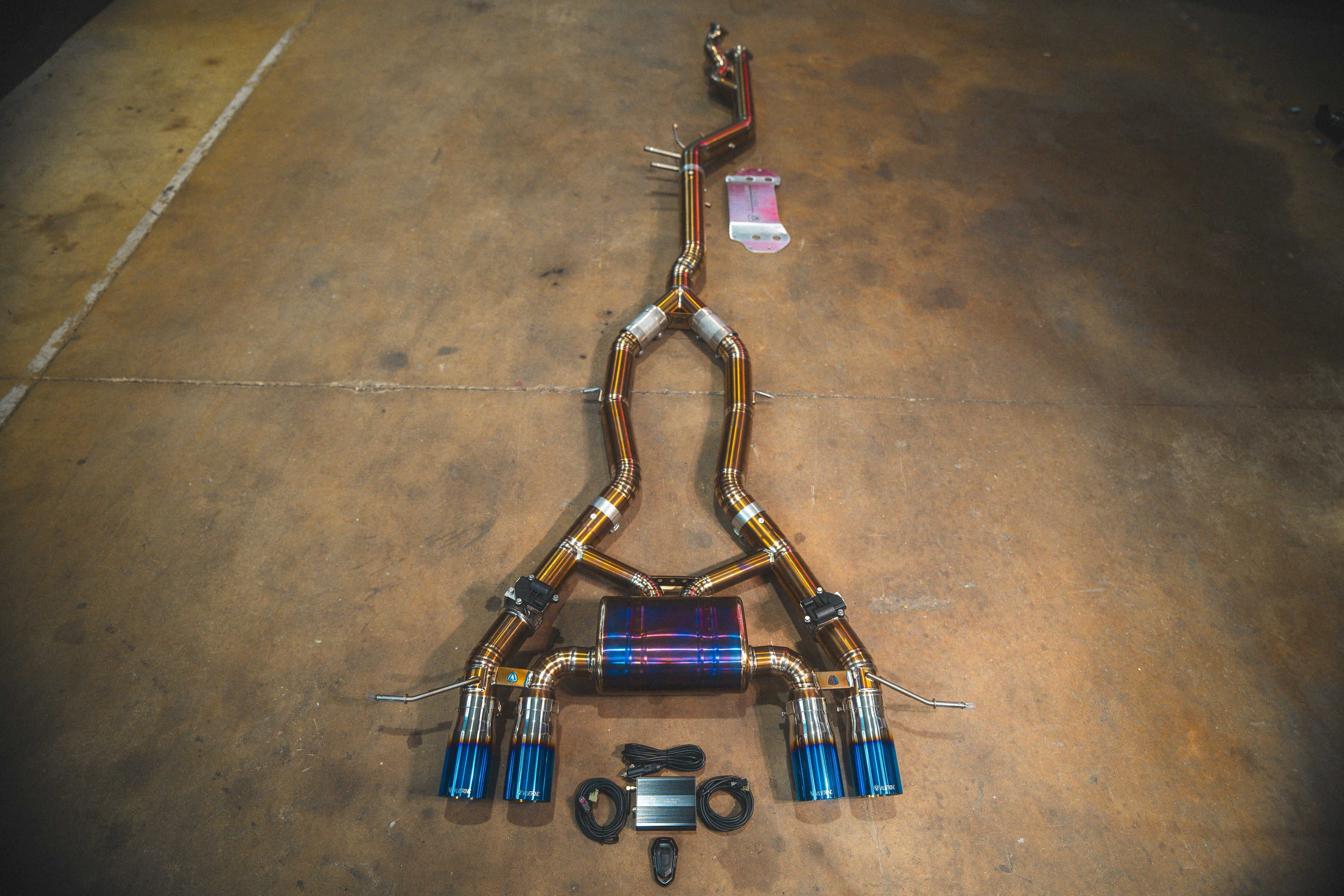 A full view of a titanium valved exhaust system for a BMW G87 M2 featuring quad exhaust tips with a blue and gold "burnt" titanium finish.