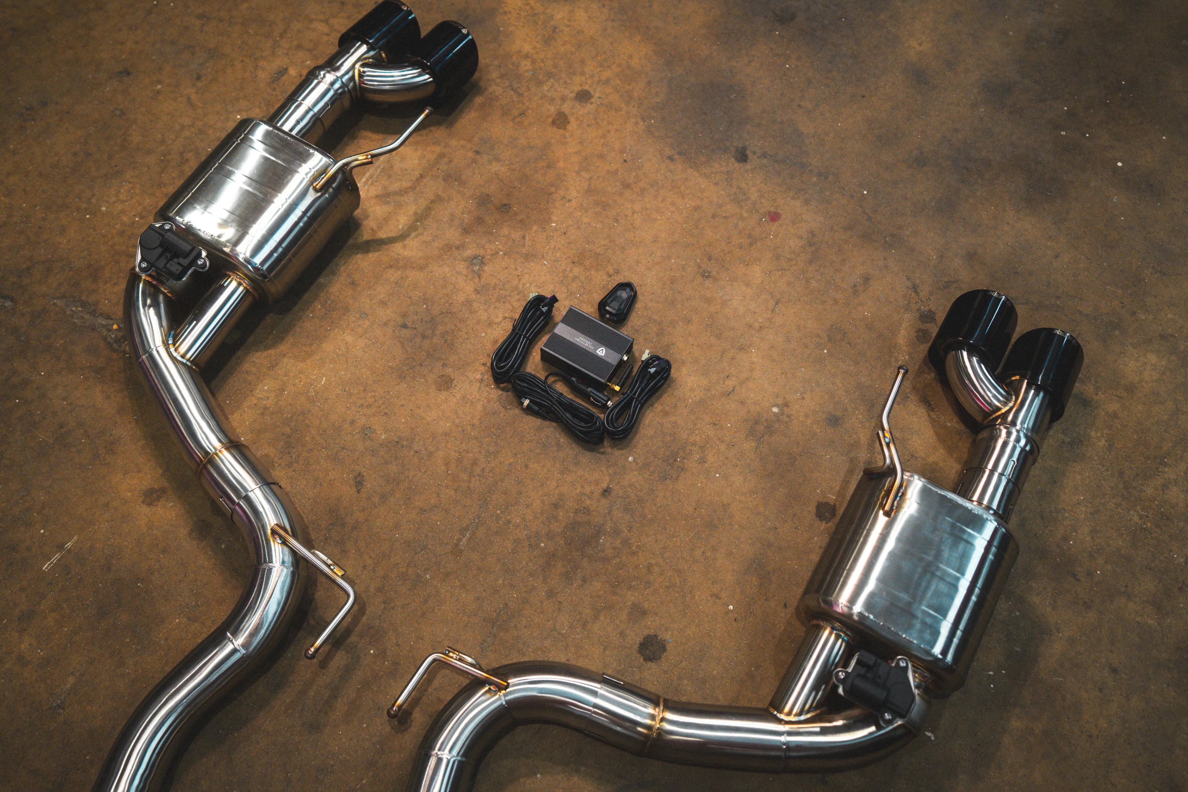 A focused view of the rear section of the exhaust system, showing the dual valved mufflers, black quad tips, and the electronic control box with wiring.