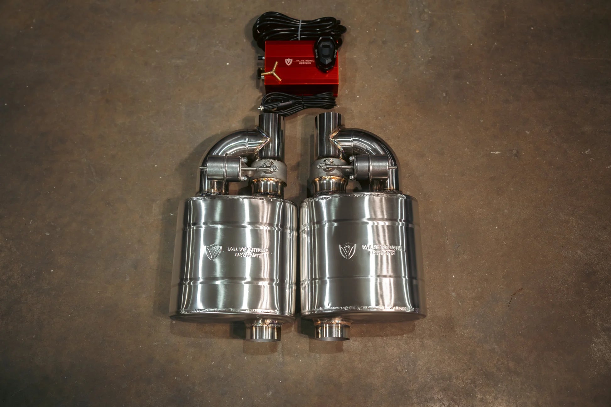 Two identical polished stainless steel mufflers with integrated valve actuators and etched logos, positioned side-by-side below a red control module and black wiring.