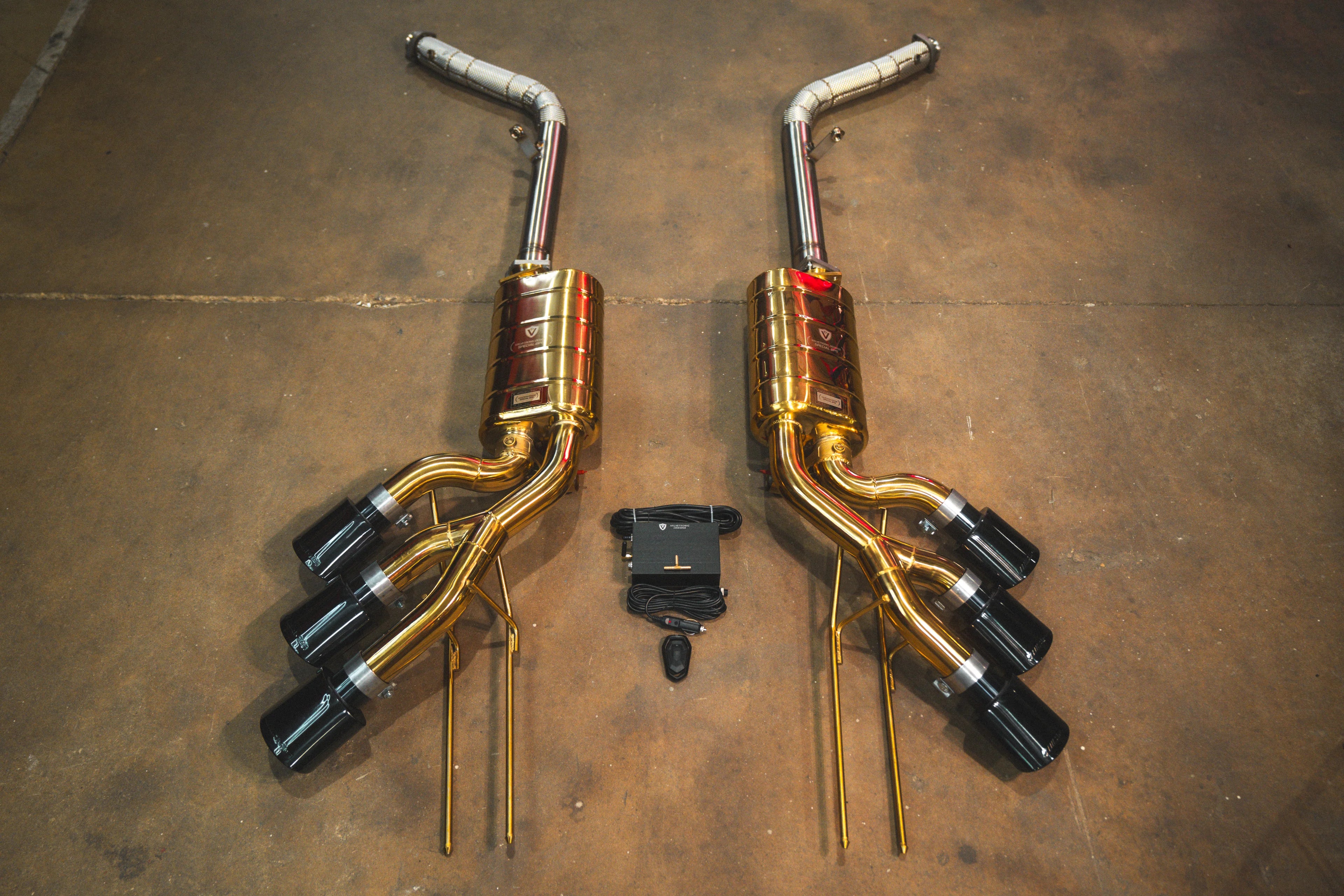 Full layout of the gold-finish exhaust system including long connecting pipes, triple-tip mufflers, and the electronic control module.
