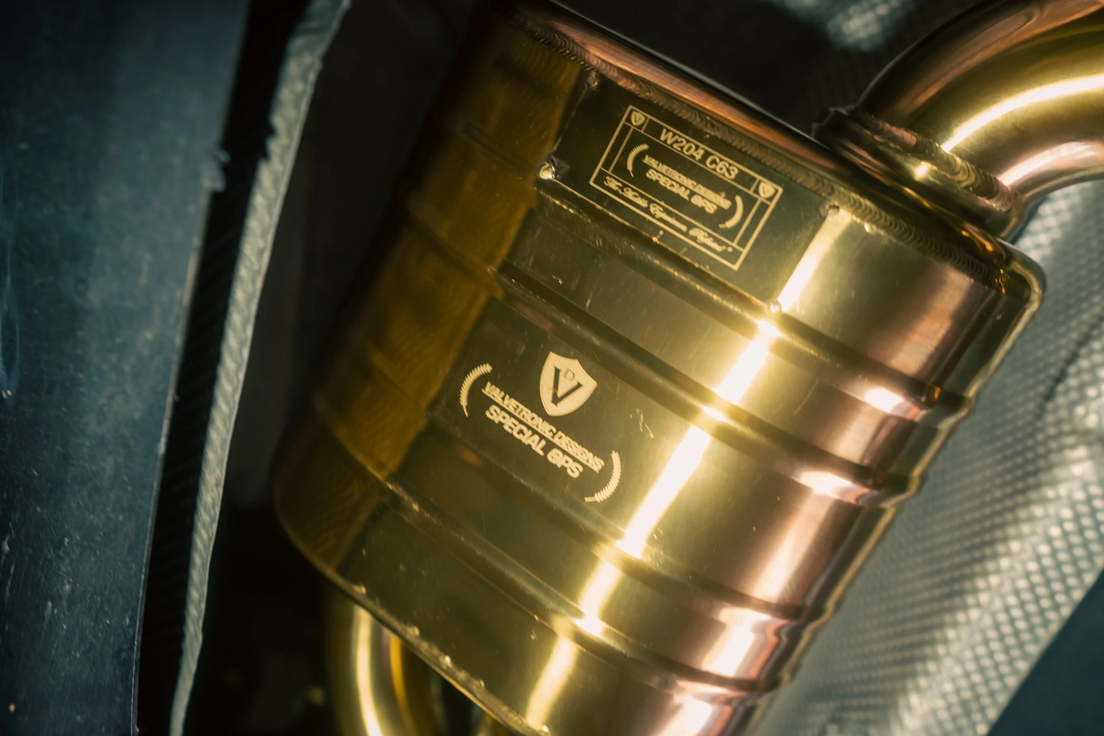 Extreme close-up of a gold exhaust muffler featuring an engraved Valvetronic Designs "Special Ops" plaque and a W204 C63 identification tag.