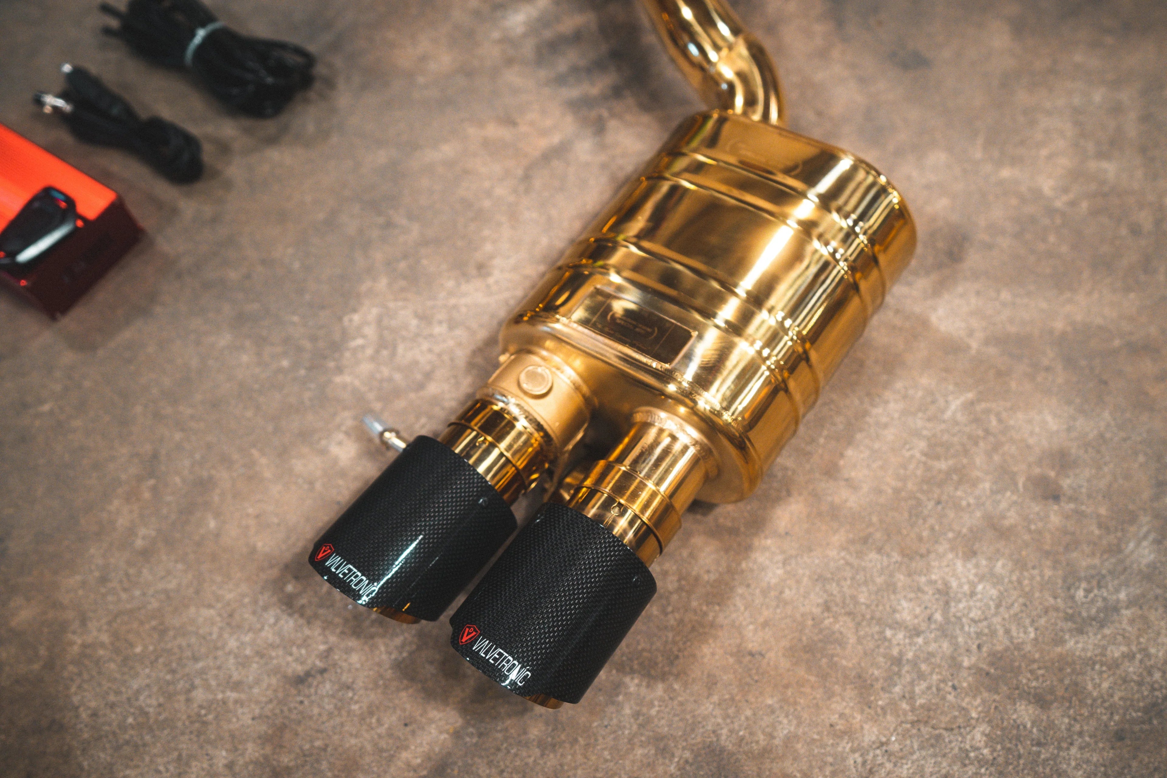 A high-angle close-up of a single anodized gold exhaust muffler connected to two matte carbon fiber exhaust tips branded with the Valvetronic logo.