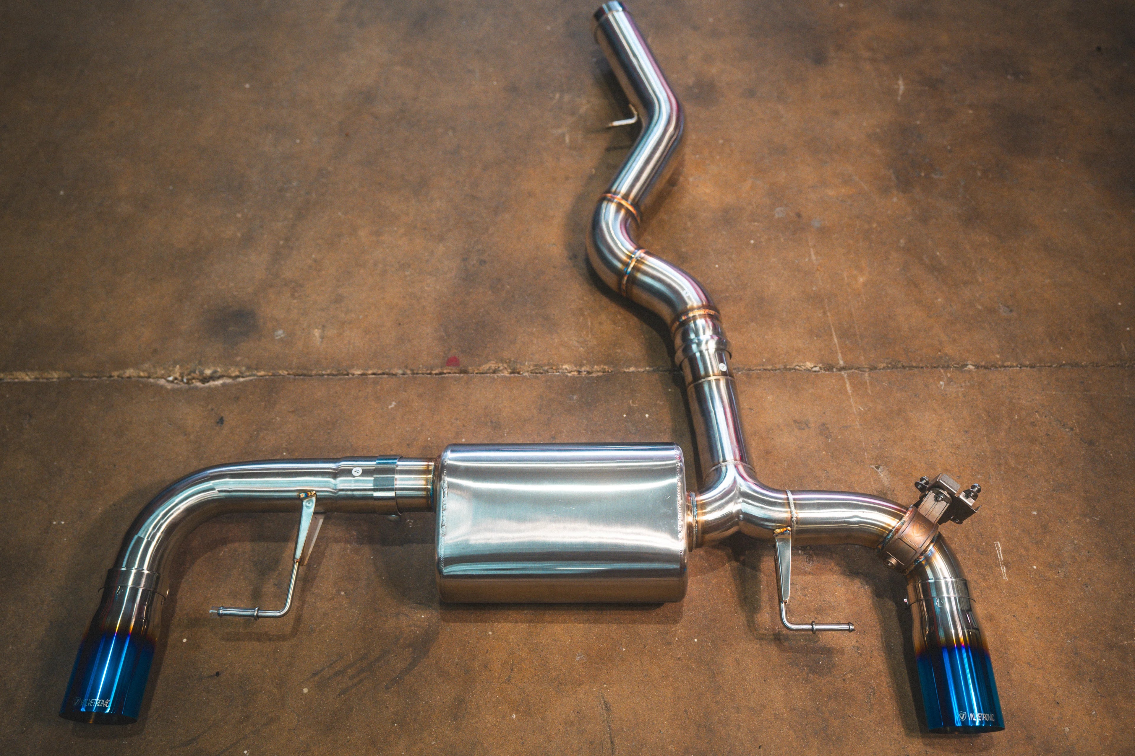 A stainless steel valved axle-back exhaust system featuring vibrant burnt blue tips resting on a brown concrete floor.