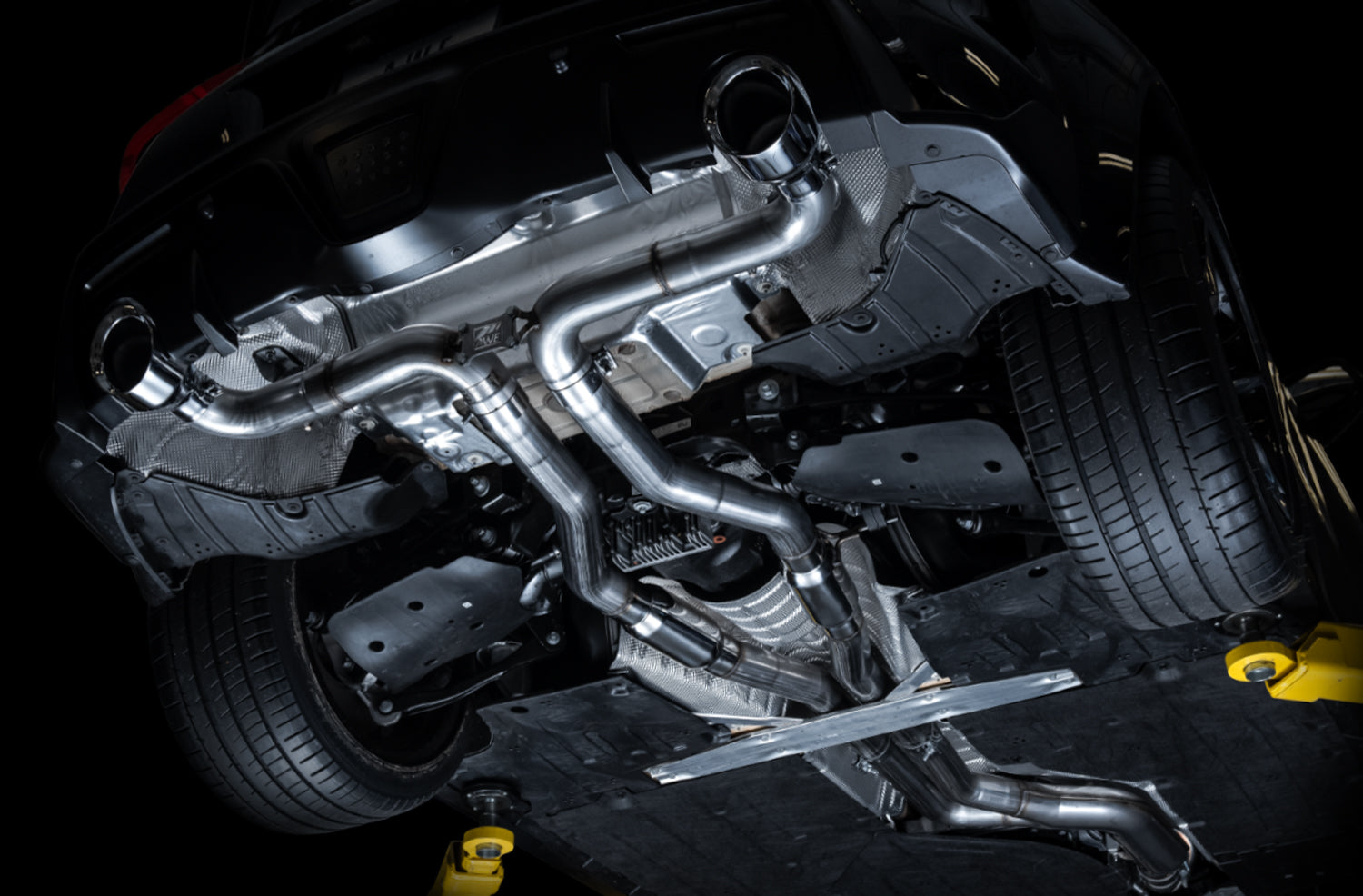 Underside of an A90 Supra on a lift showing AWE Track Edition pipework routed to the rear