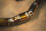 A macro close-up of a titanium pipe bend showing precise multi-pass weld beads and iridescent blue and gold heat discoloration on the metal surface.