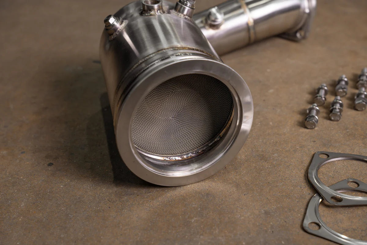A close-up view of a Valvetronic Designs 400 Cell Catted Downpipe for a 2019-2022 Lamborghini Urus, highlighting the honeycomb structure with gaskets and bolts in the background.