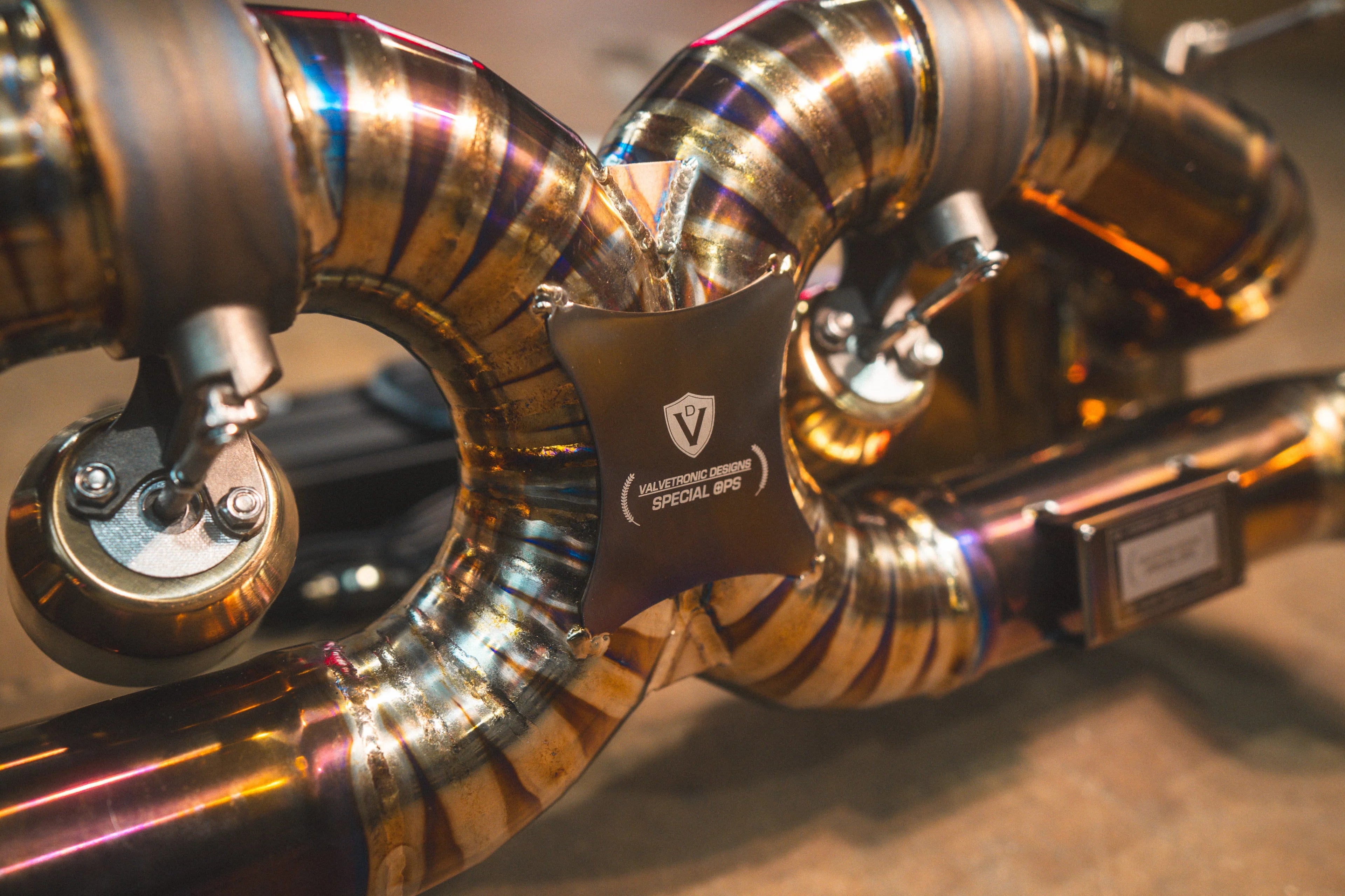 A close-up detail shot of the central X-pipe section featuring a "Valvetronic Designs Special Ops" logo plate and colorful heat-tinted welds.