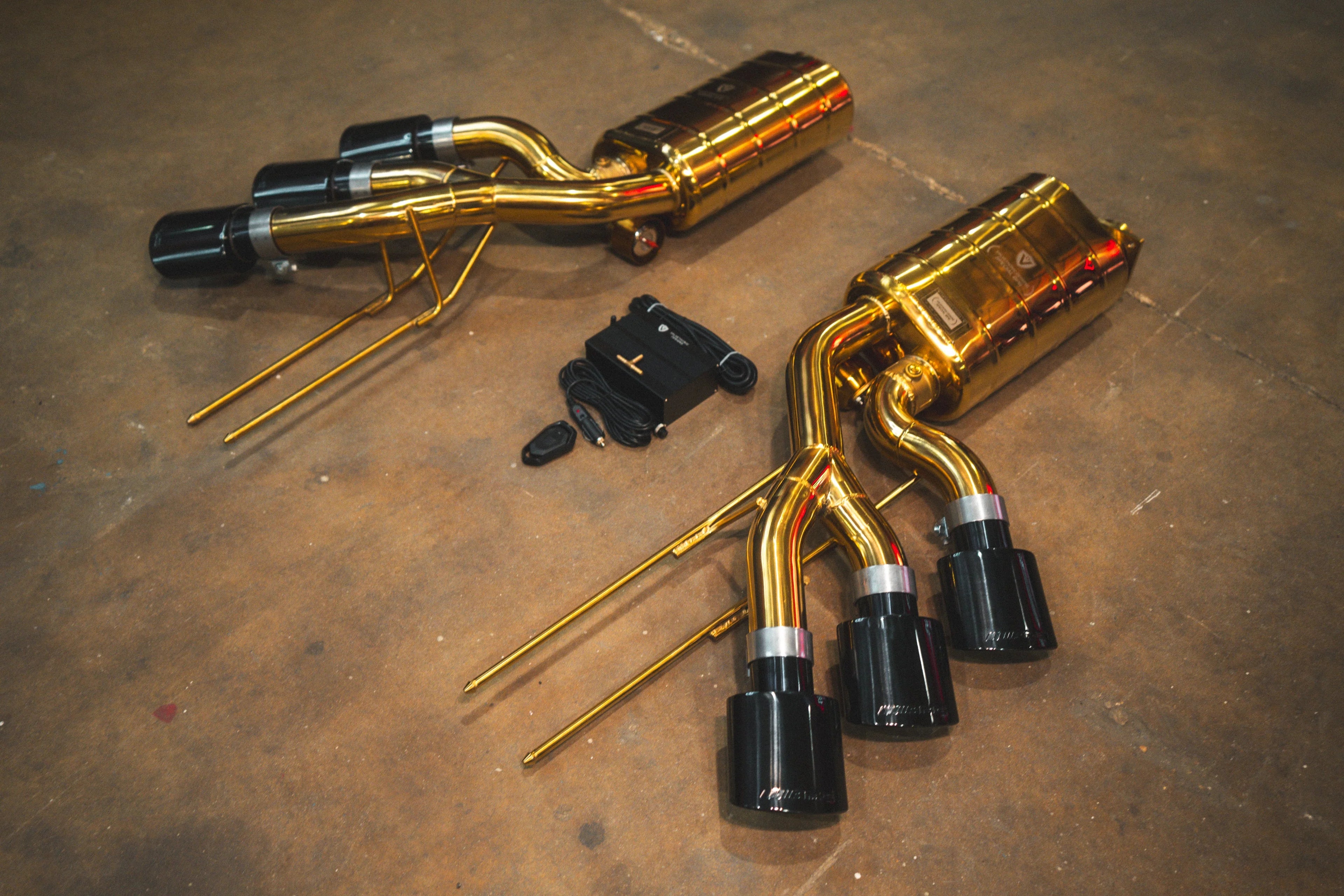 Angled perspective of the gold-plated muffler sections showing the triple black tips and gold-colored mounting hangers.