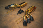 Angled perspective of the gold-plated muffler sections showing the triple black tips and gold-colored mounting hangers.