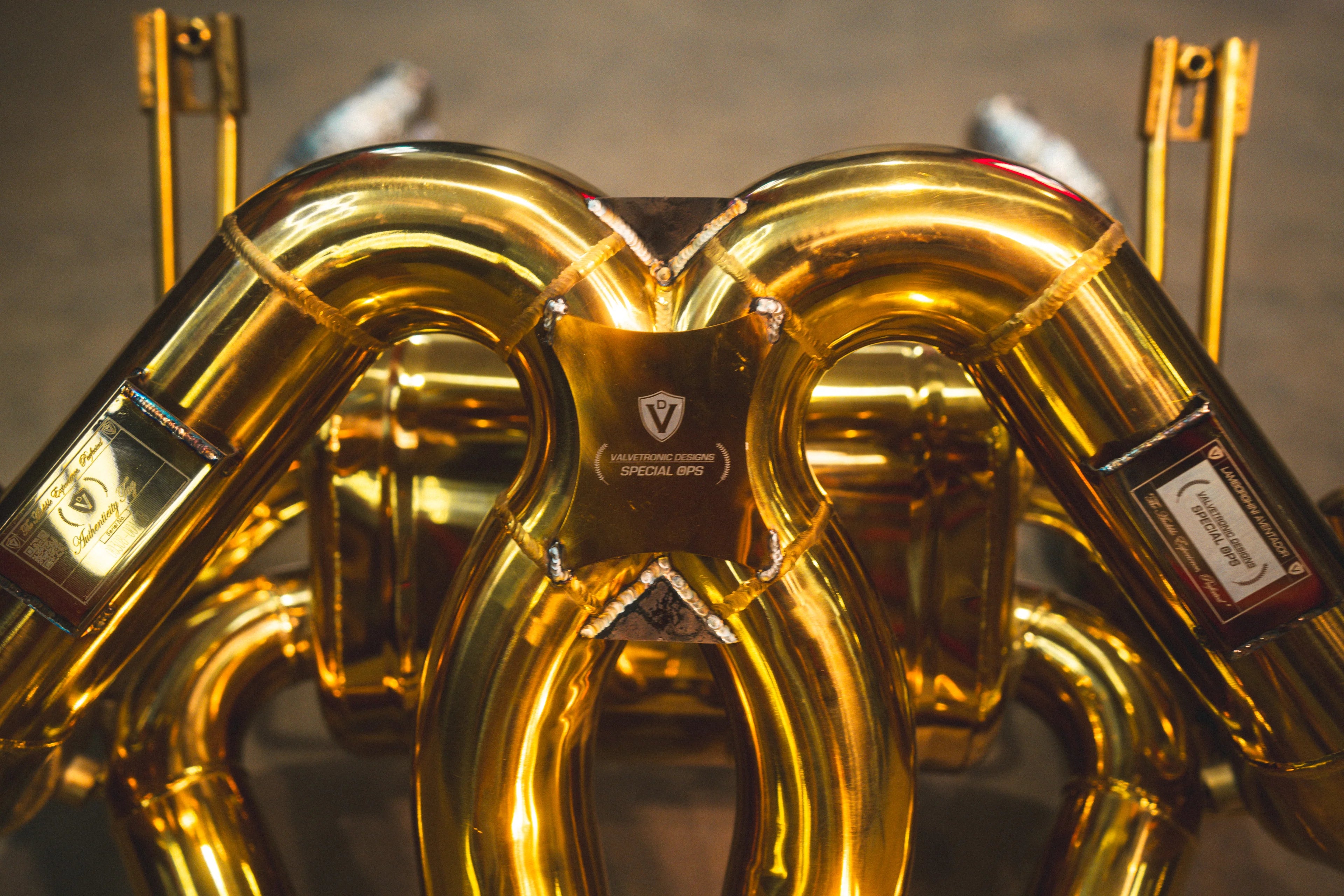 Close-up of the gold exhaust manifold center section featuring the Valvetronic Designs Special Ops logo plate and visible weld beads.