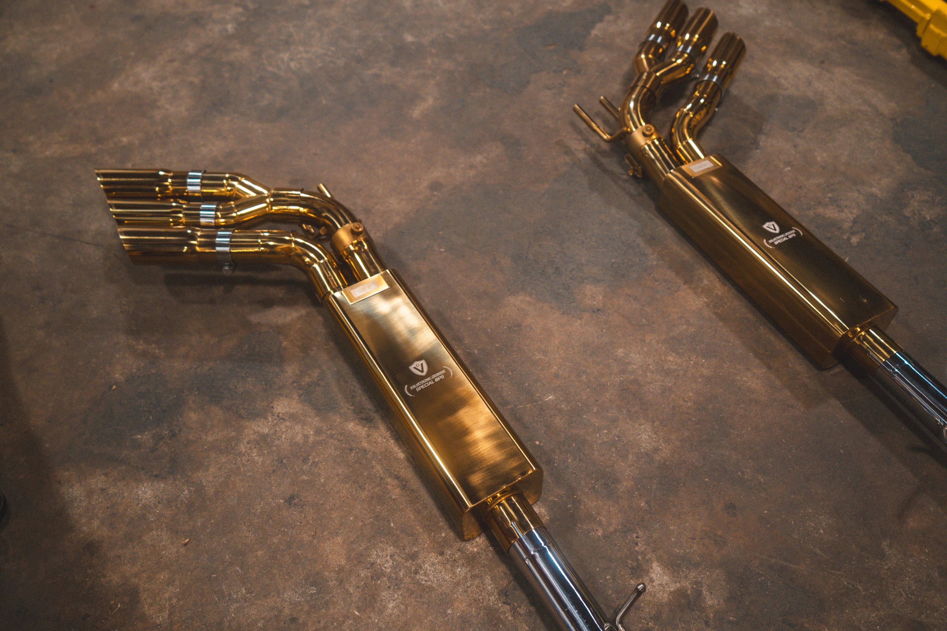 Angled perspective of the gold-finish exhaust assemblies showing the triple-tip exit and the Valvetronic Designs logo on the muffler bodies.
