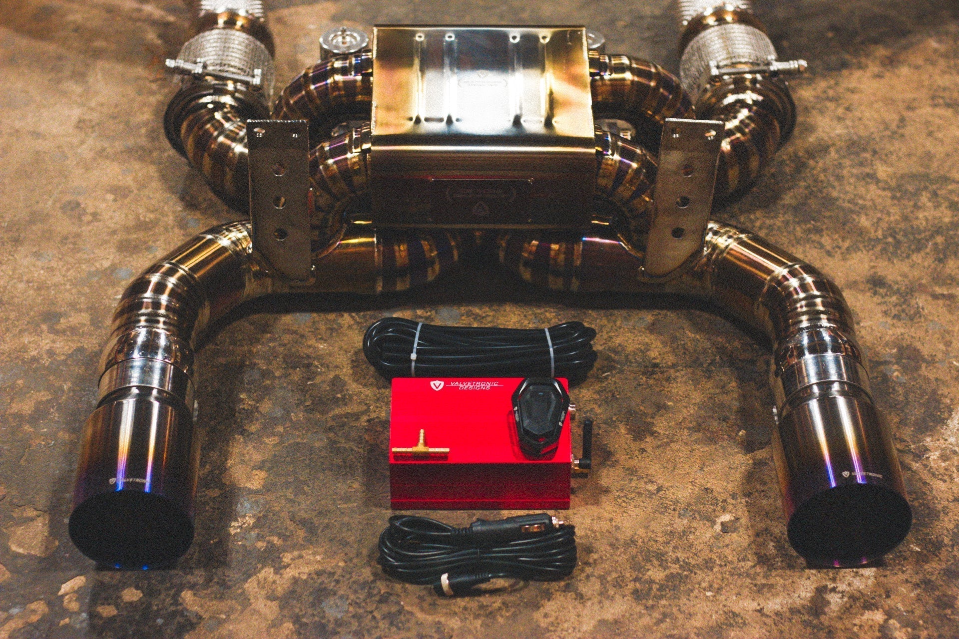 Close-up of the valved exhaust muffler assembly with a red electronic control box, remote, and wiring harness in the foreground.