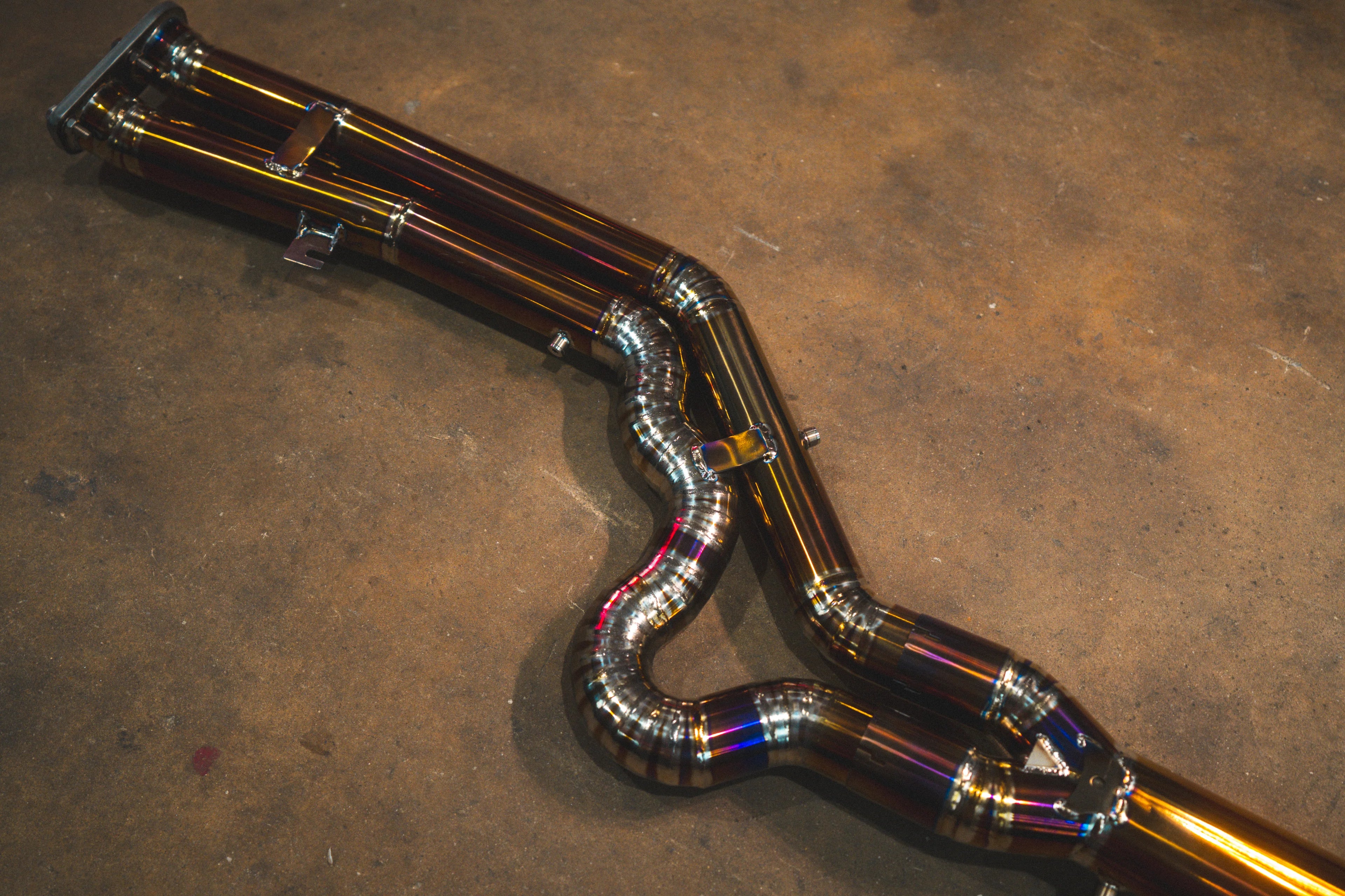 A close-up of the titanium mid-pipe section featuring a complex, snake-like bend with multiple precision-welded segments and heat-tinted coloration.