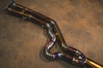 A close-up of the titanium mid-pipe section featuring a complex, snake-like bend with multiple precision-welded segments and heat-tinted coloration.