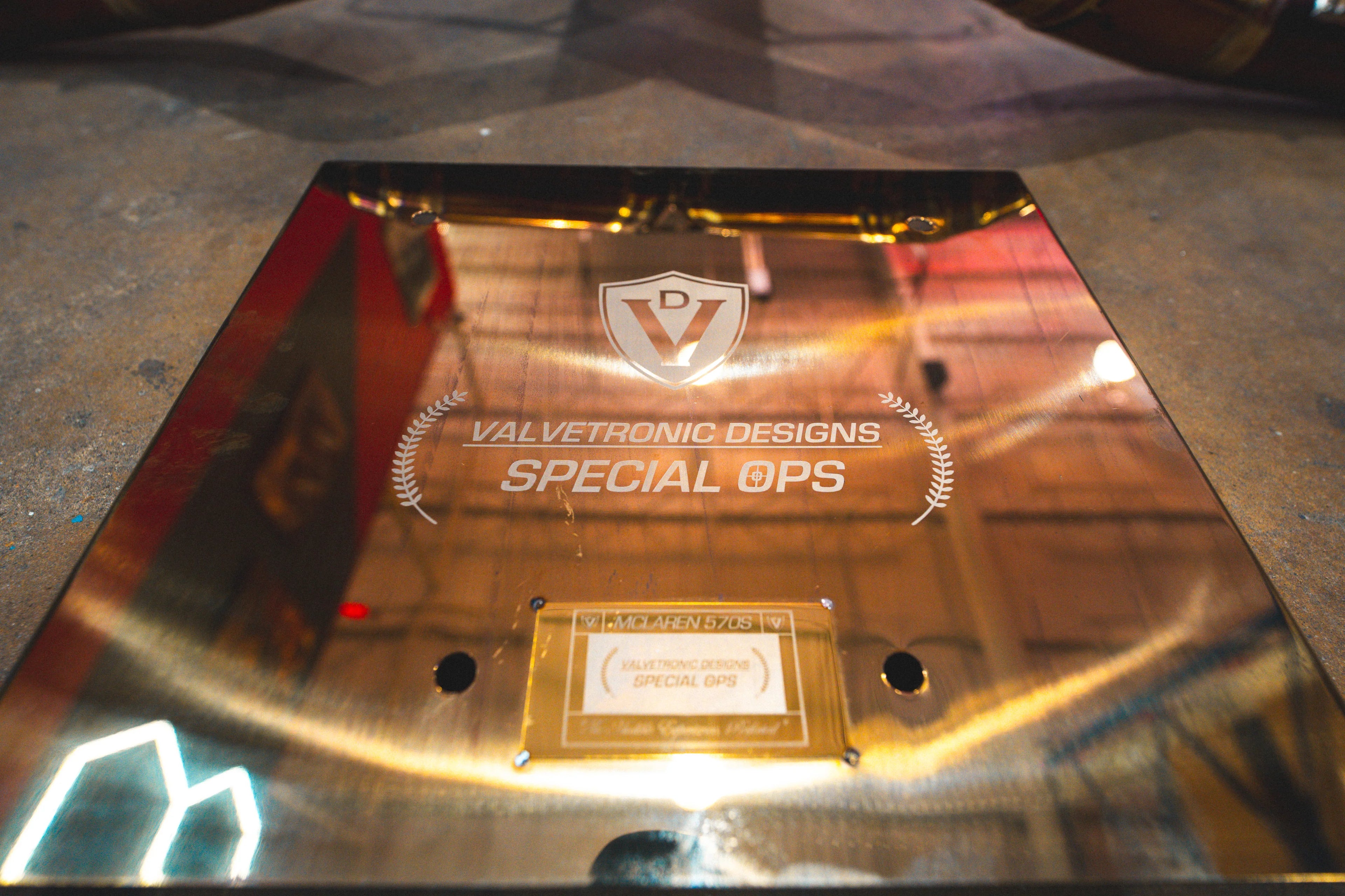 Extreme close-up of a reflective gold-finished plaque with "Valvetronic Designs Special Ops" and "McLaren 570S" engraved on it.