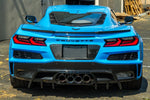 A rear three-quarter angled view of a blue Corvette C8 Z06, showcasing the carbon fiber license plate backing integrated into the rear fascia. The image highlights how the glossy carbon fiber material complements other aerodynamic components like the rear spoiler and diffuser.