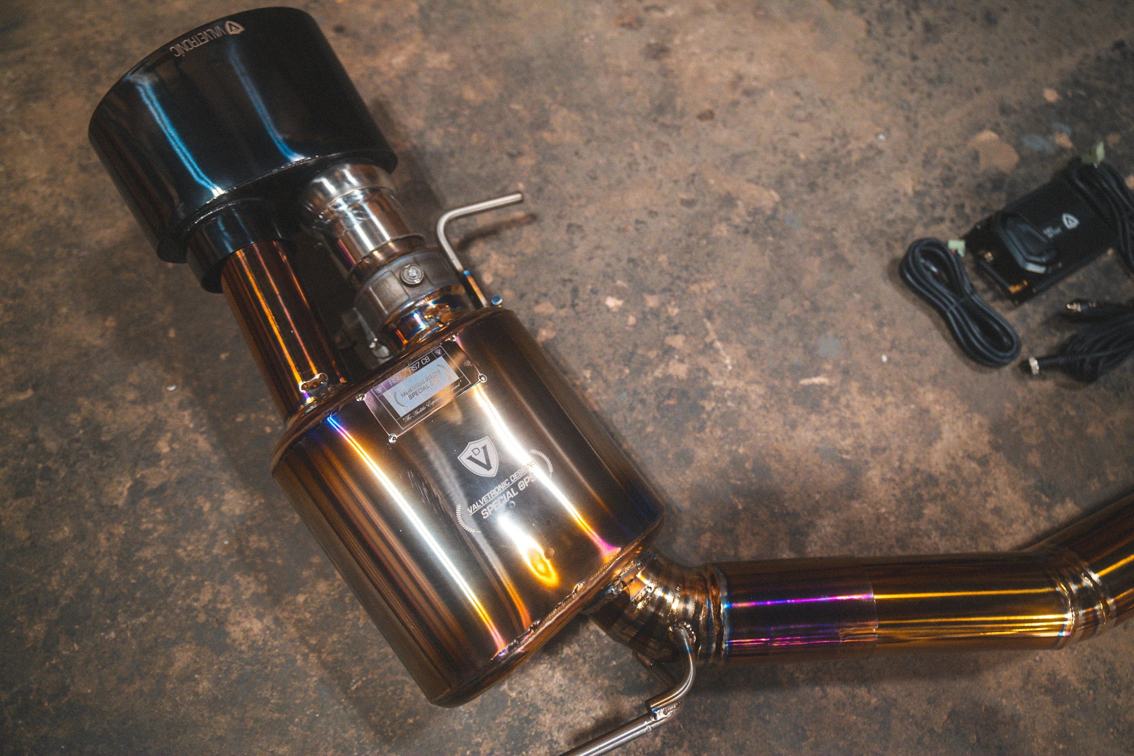 A close-up of the valved titanium muffler featuring an etched logo, connected to a large, oval black exhaust tip.