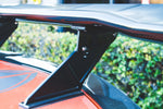 A close-up detail shot of the GTC-500 wing mounted on an orange Chevrolet Corvette C8 Z06. The photo focuses on the heavy-duty black pedestal and the adjustable mounting bracket, which allows for fine-tuning the wing's Angle of Attack (AOA). The 2x2 carbon fiber weave is clearly visible on both the wing and the spoiler delete panel.