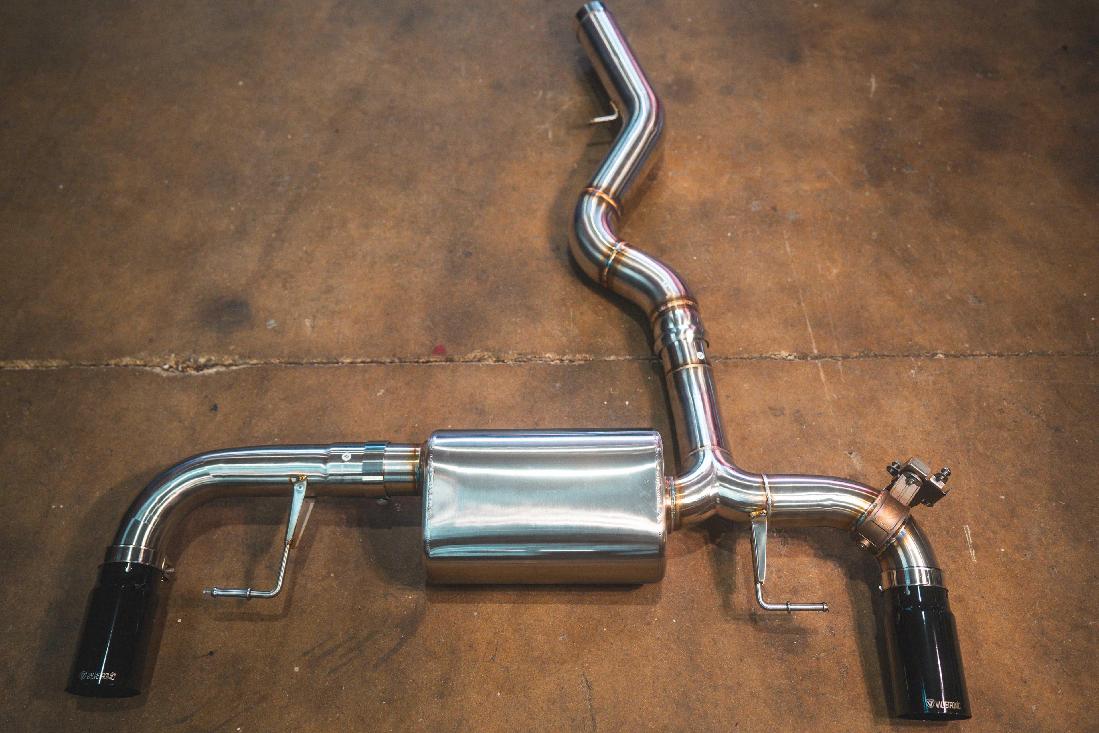 A stainless steel valved axle-back exhaust system with gloss black tips positioned on a stained concrete floor.