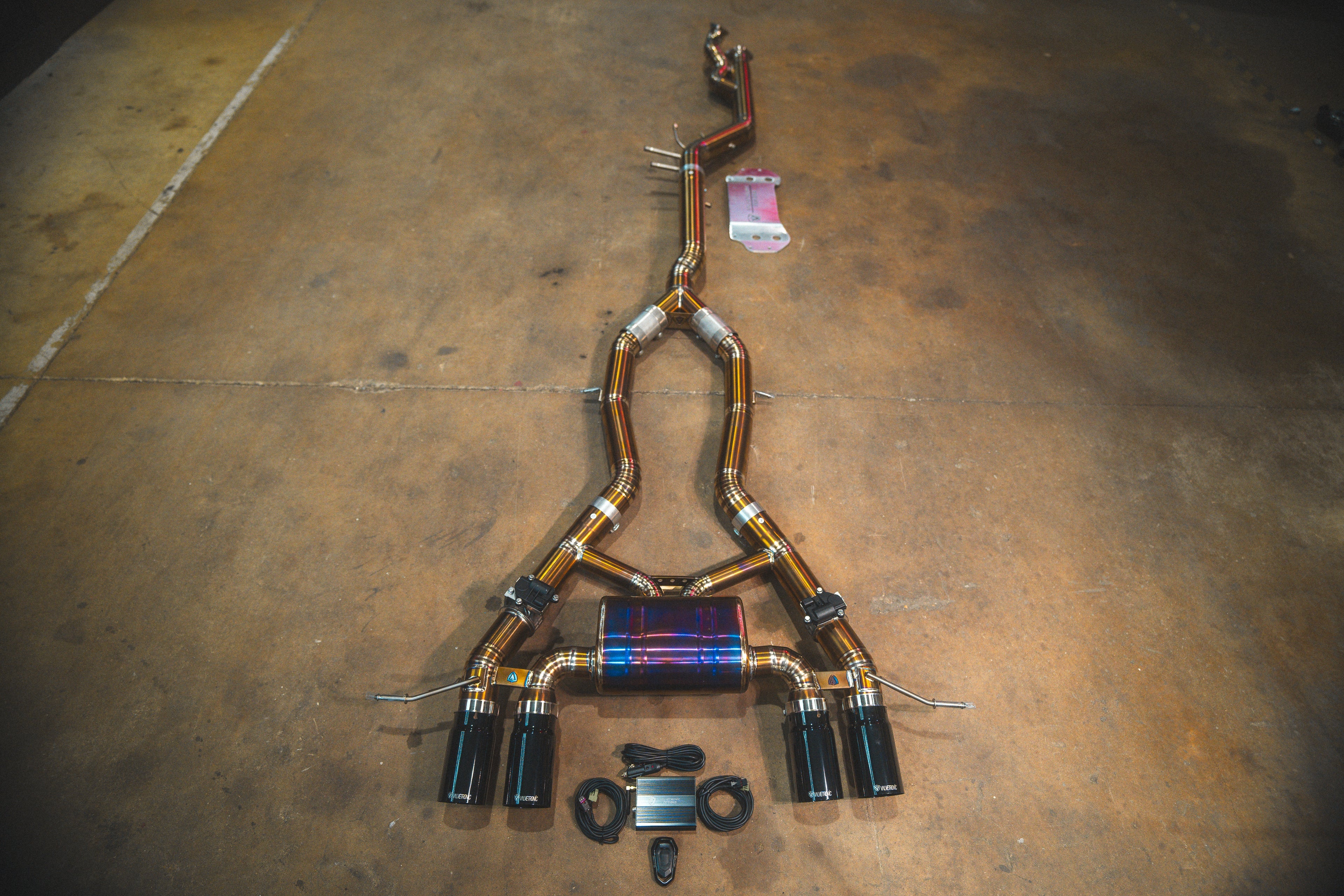 A full titanium valved exhaust system laid out on a concrete floor, featuring four gloss black exhaust tips and a central purple/blue heat-treated muffler.