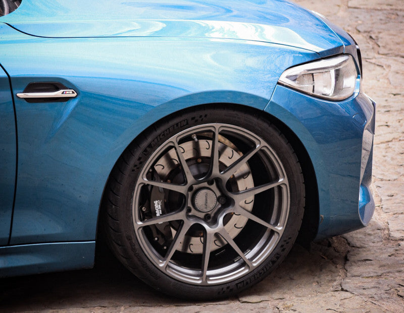 A close up view of a blue BMW with the front wheel fitted with AP Racing by Essex Radi-CAL ENP Competition Brake Kit (Front CP9660/372mm)- BMW F87 M2 & M2 Competition, F80 M3, F82 M4