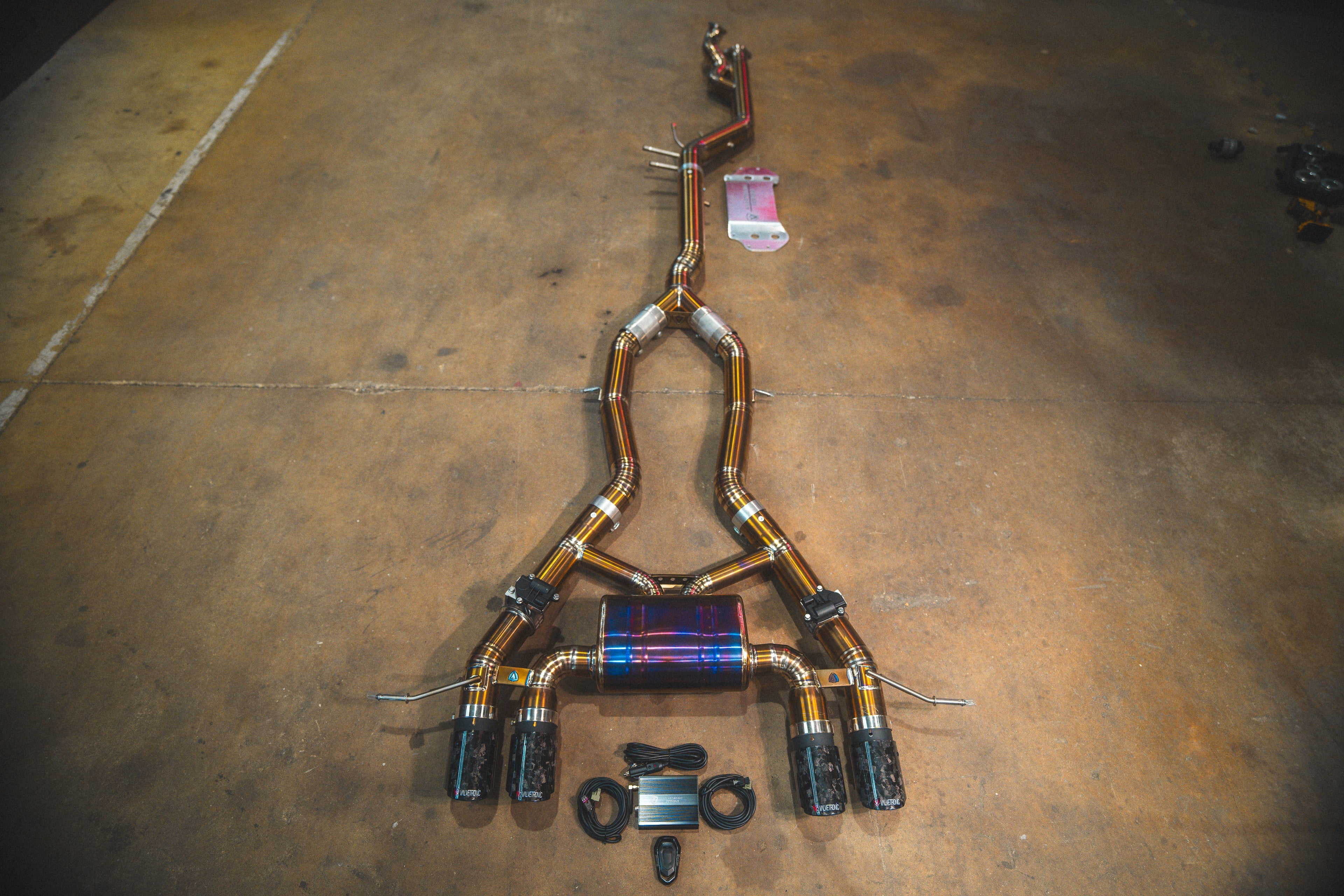 A full titanium valved exhaust system for a BMW G87 M2 featuring quad exhaust tips with a marbled forged carbon fiber texture.