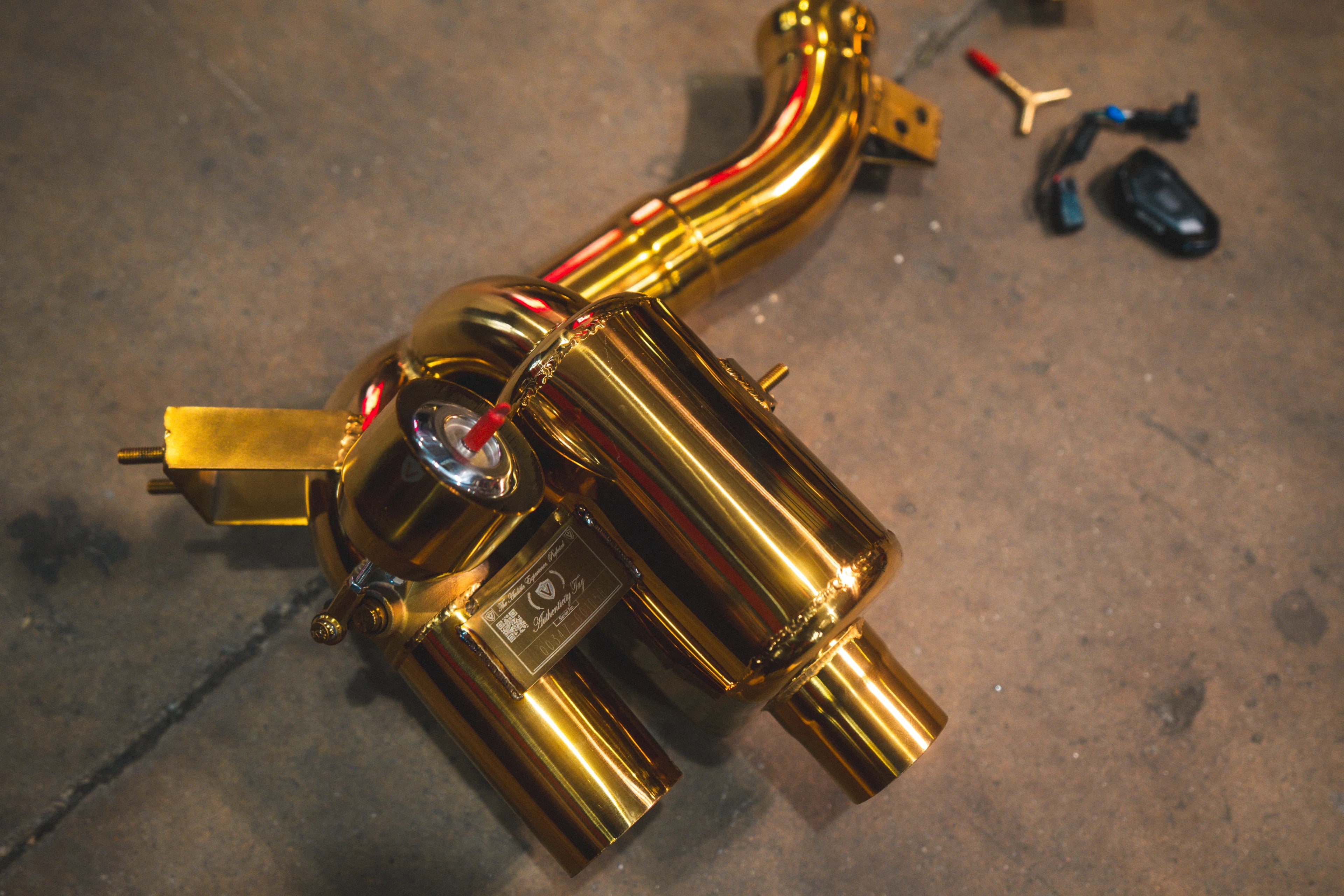 A side view of one gold-plated muffler assembly showing the vacuum-actuated valve, mounting brackets, and the authenticity plaque.