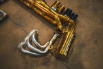 Close-up perspective of the left-side gold-finish muffler and piping assembly, highlighting the intricate pie-cut welds and the silver performance header.