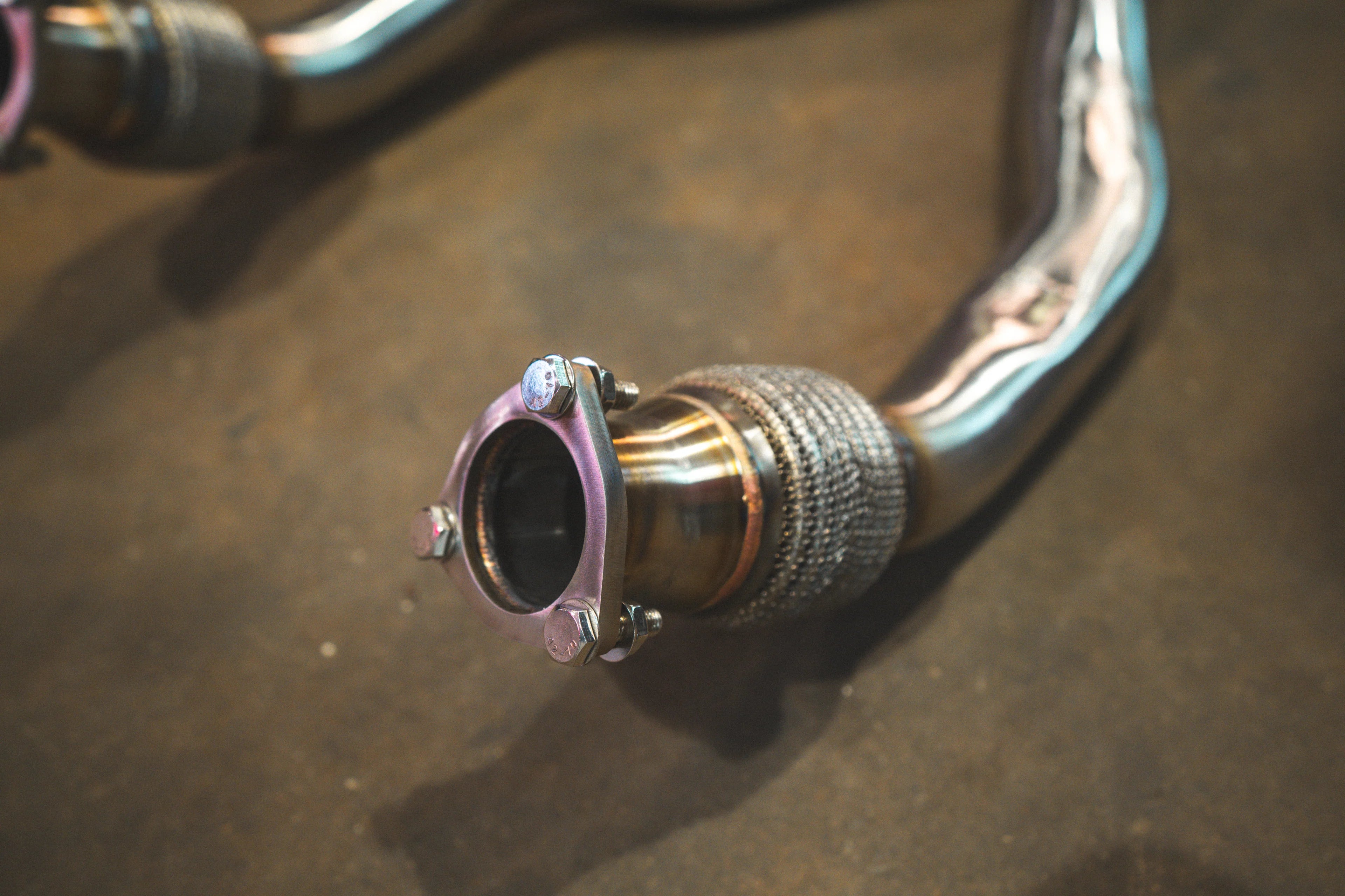 A close-up of a three-bolt exhaust flange with silver bolts secured to a titanium pipe featuring a mesh-covered flexible joint.