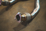 A close-up of a three-bolt exhaust flange with silver bolts secured to a titanium pipe featuring a mesh-covered flexible joint.