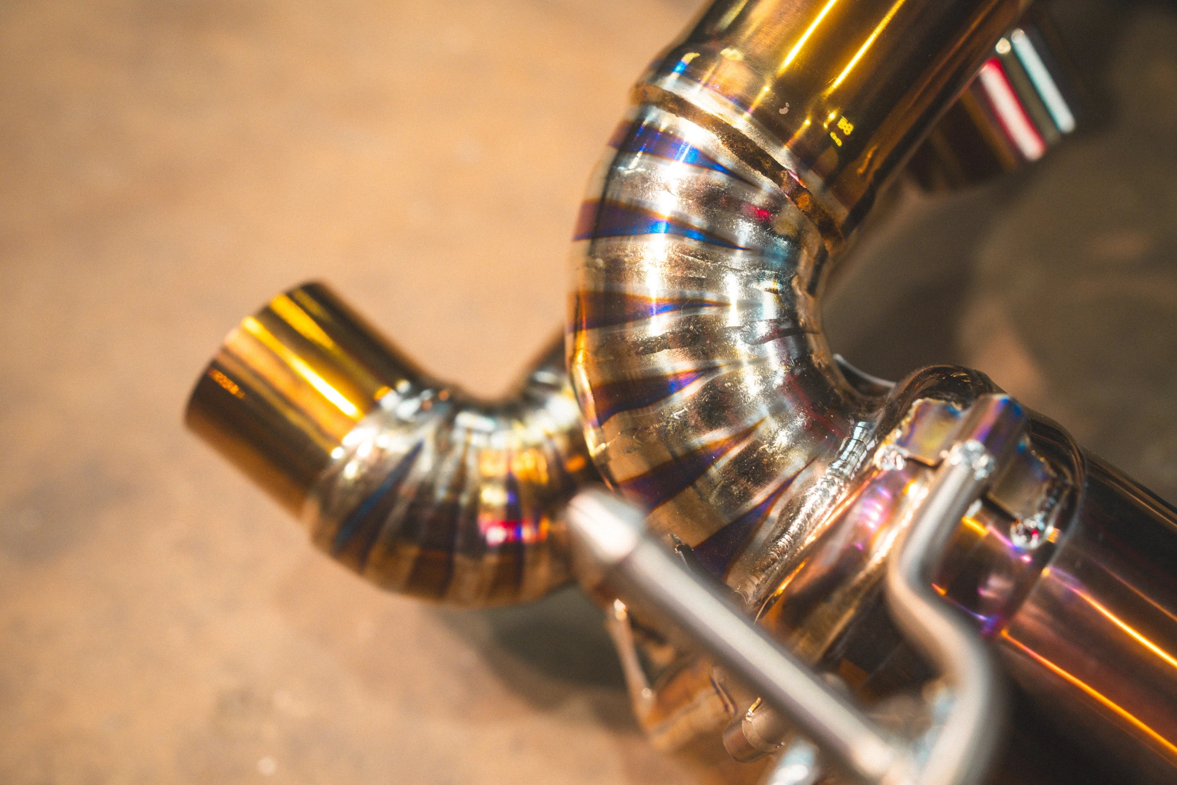 A macro view of a curved exhaust pipe section showing precision weld beads and vibrant purple and gold heat discoloration.