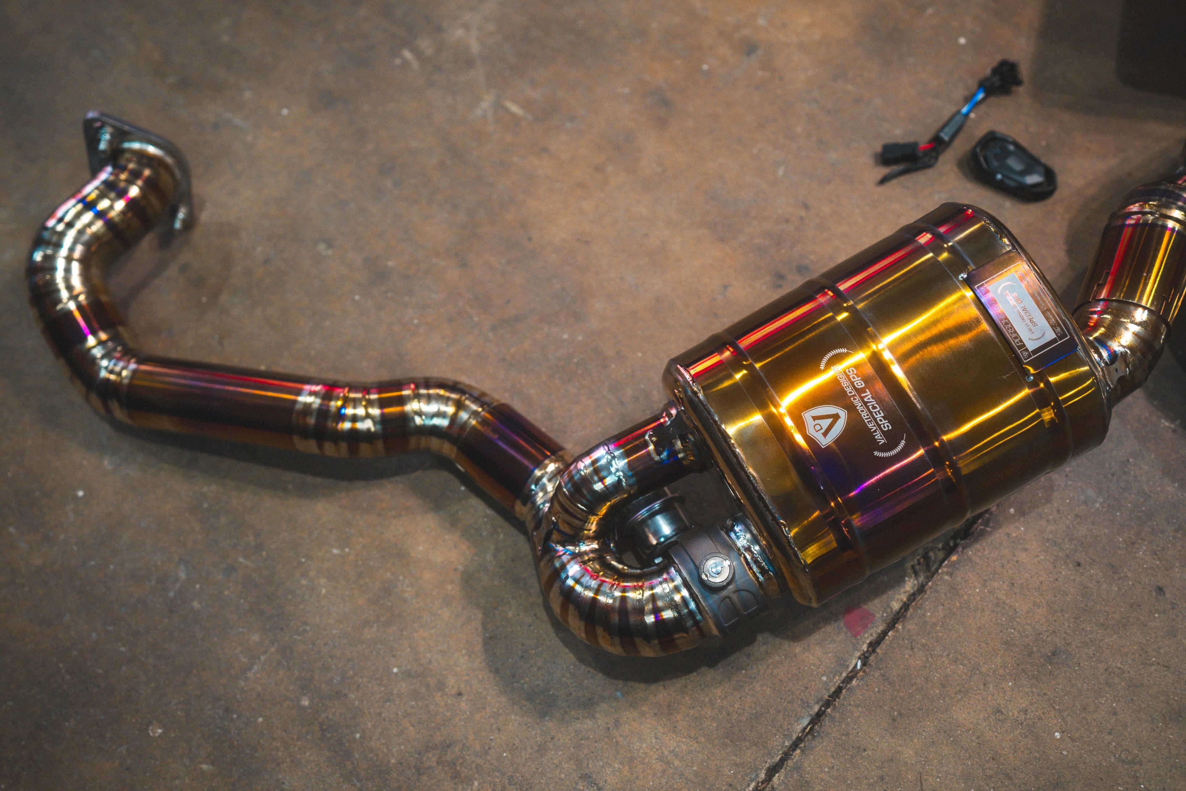Detailed view of one side of the exhaust system, showcasing the polished gold-finish muffler with "Valvetronic Designs Special Ops" branding and intricate pie-cut welds