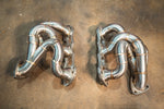 Top-down view of a pair of high-flow exhaust headers with polished piping and multi-bolt flanges.