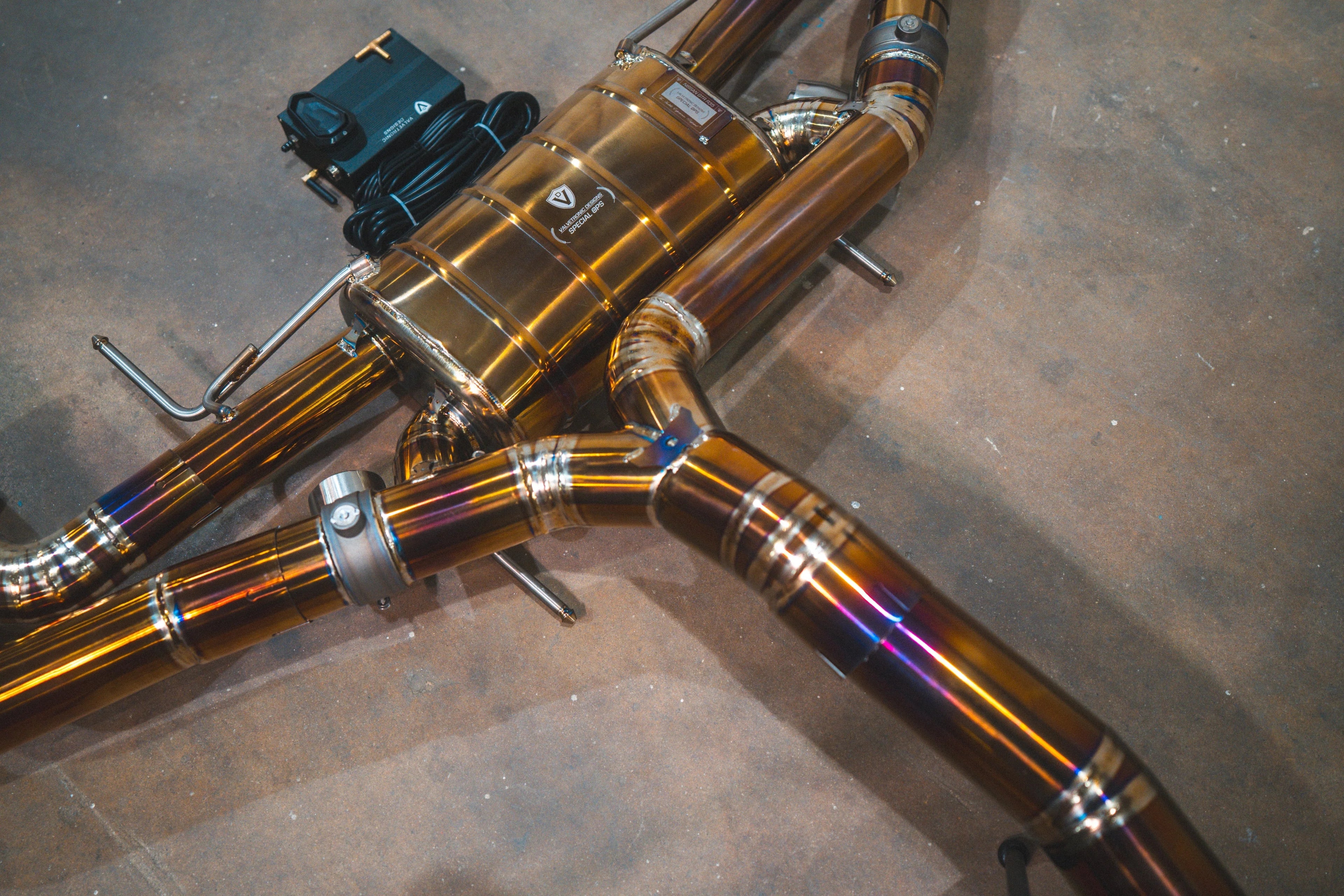 Close-up of the Y-pipe junction and center muffler, featuring the "Valvetronic Designs Special Ops" laser-etched logo on the polished gold-finish casing.