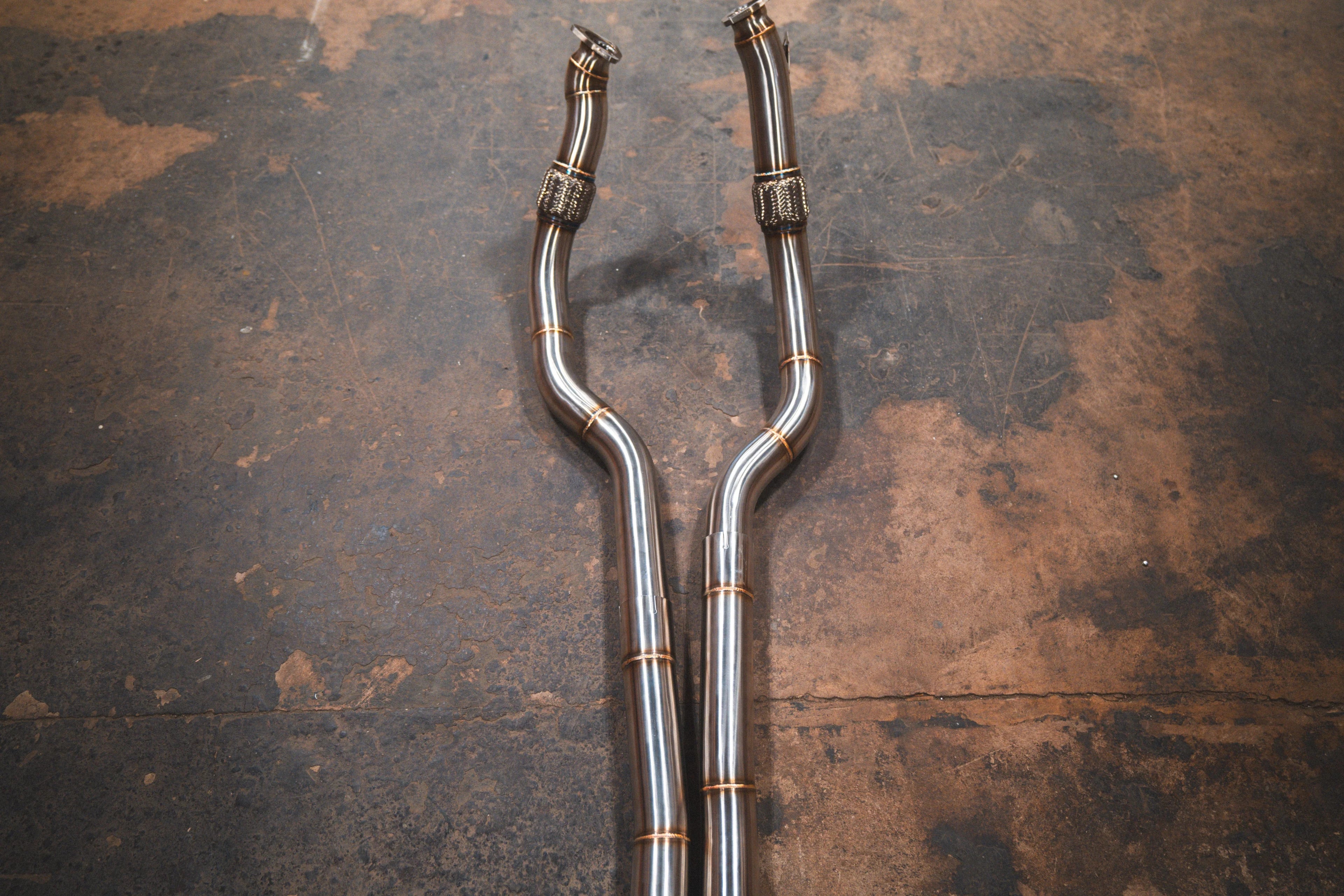 A top-down view of two polished titanium exhaust downpipes with integrated flexible mesh sections and visible weld beads.