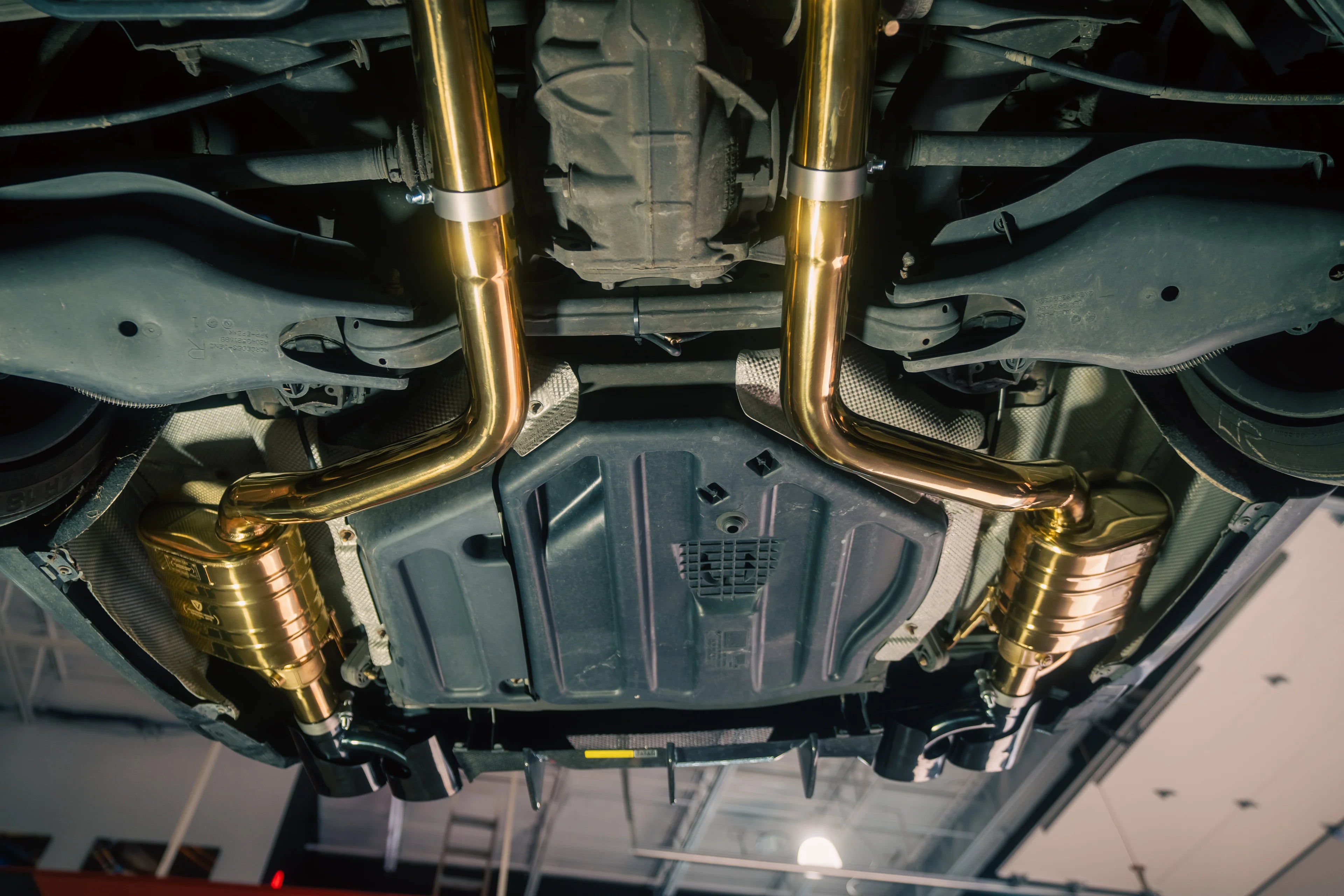 Underbody view of the full exhaust layout from the mid-section to the rear tips, showing the symmetrical gold piping and suspension components.