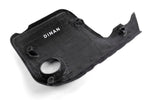 The underside of the Dinan carbon fiber engine cover, revealing the raw composite texture and integrated mounting points. The white "DINAN" brand logo is clearly visible in the center, and the circular cutout for the oil cap is positioned at the bottom.