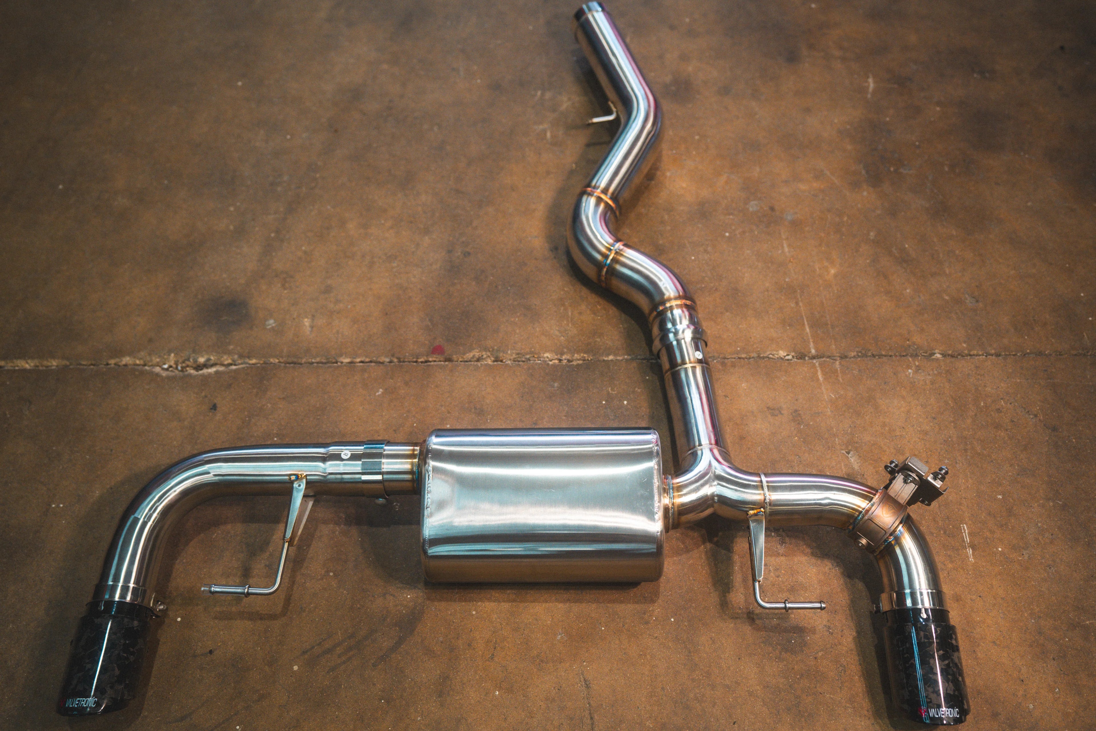 A stainless steel valved axle-back exhaust system with marbled forged carbon fiber tips on a brown concrete floor.