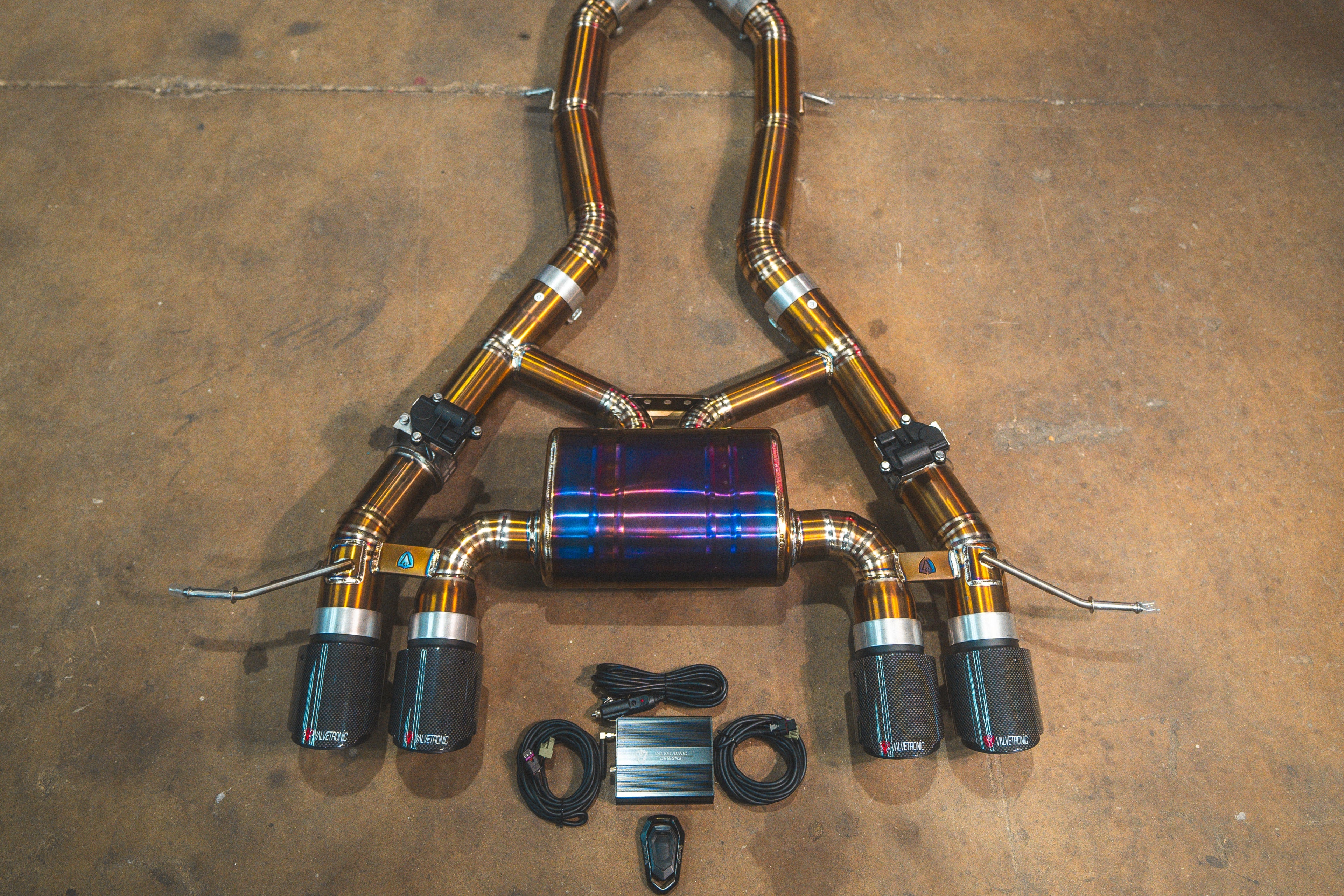 A close up view of the titanium axle back valved exhaust system for a BMW G87 M2 featuring quad outlets with traditional weave carbon fiber exhaust tips