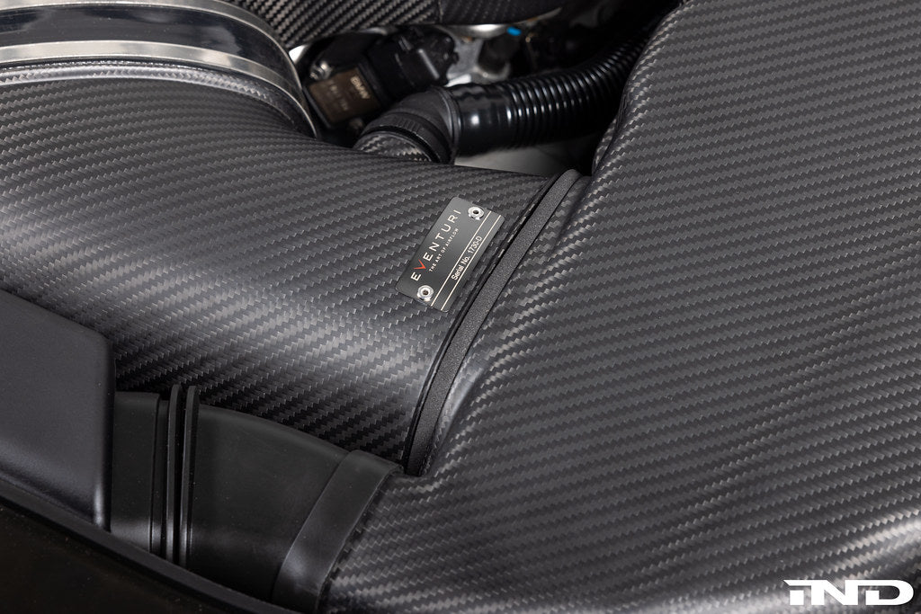 Carbon fiber intake box with label installed in engine bay showing filter housing and connecting hoses - Eventuri cold air intake for BMW M3